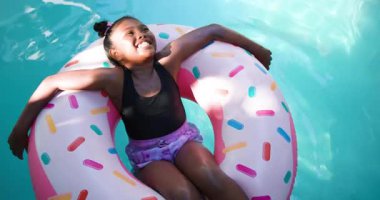 Çocuk kız yüzen kenarları itiyor, turkuaz havuz suyunda donut döndürüyor ve yukarı doğru gülümsüyor. Oyunculuk, rahatlama, rahatlama, yaz tatili, açık hava, mutluluk, coşku