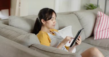 Kanepeye kurulduktan sonra Asyalı kadın siyah tableti tıklatıp eğlenceli bir yazı okuduktan ve mutlu bir şekilde esnedikten sonra. Rahatlama, rahatlama, teknoloji, konfor, rahatlık, rahatlık, ev