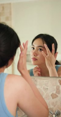 Dikey video: Genç Asyalı kadın banyo aynasında derisini inceliyor, cilt bakımına odaklanıyor. Güzellik, sağlık, kendine bakım, kozmetik, yansıma, hijyen