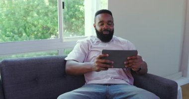 Modern oturma odasındaki gri koltukta oturan Afrikalı Amerikalı adam tablet ve fotokopi alanına odaklanıyor. Rahatlama, teknoloji, modern, rahat yaşam tarzı, odaklanma, yeşillik