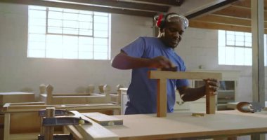 Parlak atölyedeki kaba noktayı işaret eden iş arkadaşından sonra Afro-Amerikalı bir adam T şeklindeki çerçeveyi zımparalıyor. Sanatçı, marangozluk, marangozluk, atölye, işçilik, diy, yenileme