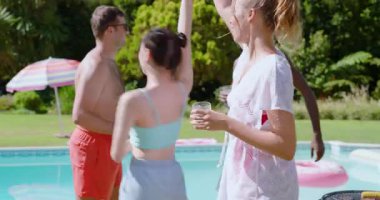 Farklı arkadaşlar kadehleri kaldırıyor pembe örtülü kadın gökyüzünü işaret ediyor, havuz ızgarasının etrafında dans ediyor. Kutlama, eğlence, açık hava, yaz, arkadaşlık, neşe, sükunet