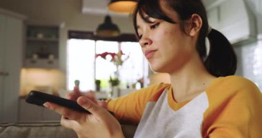 Asyalı kadın ihbardan sonra telefonu kaydırıyor, hoparlör moduna geçiyor ve kanepede gülümsüyor. Teknoloji, iletişim, yaşam tarzı, ev, bağlantı, modern, etkileşim