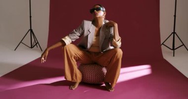 Noticing light pattern, Indian woman in beige blazer adjusting sunglasses, posing on pouf in studio. Fashion, elegance, glamour, contemporary, portrait, studio, confidence