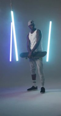 Dikey video: Genç bir adam modern stüdyoda neon ışıklı kaykay tutuyor. Kaykay, güven, şehir, moda, moda, gençlik