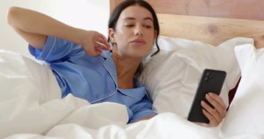 Mavi pijamalı bir kadın evde akıllı telefon kullanırken gülüyor. Teknoloji, dinlenme, konforlu, dijital, yaşam tarzı, boş zaman