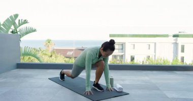 Yetişkin bir Asyalı kadın balkona çıkıyor, esnemek için yeşil su şişesinin yanında hasırını açıyor. Spor, sağlık, açık hava, sükunet, dinginlik, egzersiz, sağlık