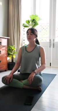 Dikey video: Asyalı kadın evde yoga minderi üzerinde meditasyon yapıyor, farkındalığa odaklanıyor. Meditasyon, rahatlama, sükunet, sağlık, zen, dinginlik