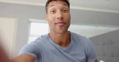 Yetişkin bir adam ileri uzanıyor akıllı telefon kamerasını ayarlıyor ve modern yatak odasında lense gülümsüyor. Portre, vlogger, modern iç mekan, samimi, kendinden emin, anlamlı, yaşam tarzı