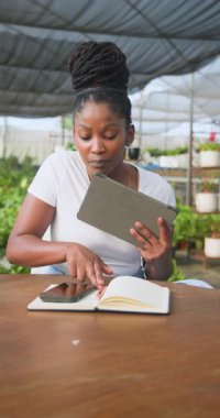 Dikey video: Afro-Amerikan kadın serada tablet tutarken deftere yazı yazıyor. Bahçıvanlık, bahçıvanlık, teknoloji, dijital, tarım, sürdürülebilirlik