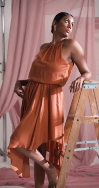 Dikey video: Stüdyodaki ahşap merdivende güvenle poz veren zarif turuncu elbiseli model. Moda, zarafet, portre, tarz, güzellik, modellik