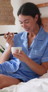 Dikey video: Mavi pijamalı gülümseyen kadın kahve içiyor ve yatakta akıllı telefon kullanıyor. Sabah, rahatlama, teknoloji, iletişim, rahatlık, rahatlık