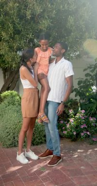 Dikey video: Güler yüzlü aile açık havada güneşli bir günün tadını çıkarıyor, ebeveynler kızını bahçede tutuyor. Mutluluk, kaynaşma, boş zaman, yaz zamanı, birliktelik, kaygısızlık