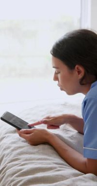 Dikey video: Mavi üniformalı genç bir kadın yatakta dinleniyor, tablet okurken gülümsüyor. Teknoloji, rahatlama, yaşam tarzı, ev, boş zaman, dijital