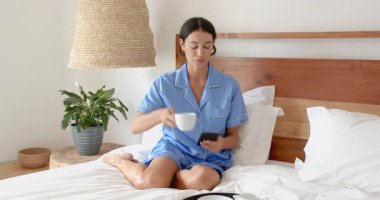 Yatakta dinlenen, mavi pijamalı bir kadın sabah kahvesinin tadını çıkarıyor, evinde mutlu mesut gülümsüyor. Rahatlama, konfor, yaşam tarzı, rahatlık, rahatlık, mutluluk