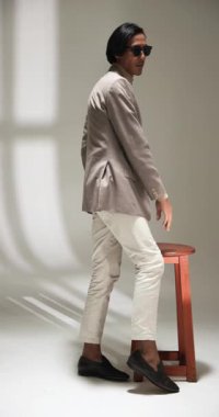 Dikey video: Güneş gözlüklü şık adam stüdyoda ahşap tabureye ceket ayarlıyor. Moda, erkek giyim, tarz, moda, göz giyimi, özgüven