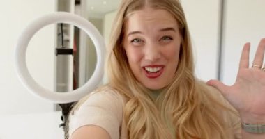 Telefonları dinlemek, seyircilere el sallamak ve teşekkür etmek, yatak odası masasında gülümseyen kadın video kaydedicisi, halka ışığı altında. Kişisel markalaşma, sosyal medya, nüfuz, içerik oluşturma, minimalist, canlı, yaşam tarzı