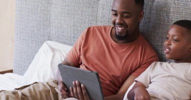 Afro-Amerikalı baba ve oğulun tablete dokunması oğlunun içerik solosunu yatakta göstermesini tetikliyor. Ebeveynlik, eğitim, teknoloji, bağlanma, gevşeme, modern, destekleyici