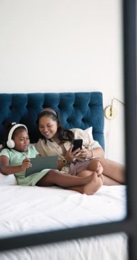 Vertical video: Mother and daughter relaxing on bed, using smartphone and tablet together. Family, bonding, technology, leisure, lifestyle, digital