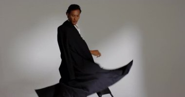 Asian male holding coat beside chair, lifting coat for drama then walking and sitting in studio. Fashion, elegance, versatility, portrait, monochrome, style, sophistication