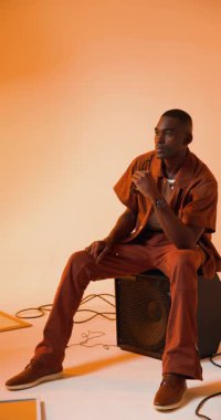 Dikey video: Genç adam şık bir stüdyoda hoparlörde otururken güneş gözlüğünü ayarlıyor. Moda, tarz, müzik, trend, ses, eğlence