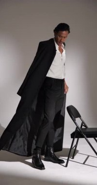 Dikey video: Moda stüdyosunda kendine güvenen şık takım elbiseli bir adam sandalyeye oturuyor. stil, özgüven, erkek giyim, zarafet, fotoğraf, poz