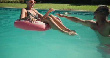 With man giving gentle push, senior couple spinning on pink donut float in backyard pool laughing. Leisure, recreation, summer, playful, outdoor, companionship, vibrant