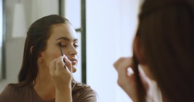 Female wearing brown top glancing at mirror, using eyeshadow brush at vanity and applying lip gloss. Beauty, self-care, grooming, elegance, sophistication, femininity, lifestyle