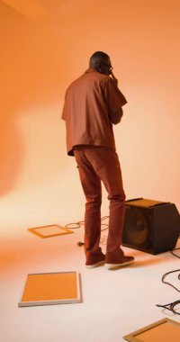 Dikey video: şık bir stüdyoda hoparlörde oturan, güneş gözlüğü takan Afrikalı Amerikalı bir adam. Portre, çağdaş, müzik, yaşam tarzı, moda, kentsel