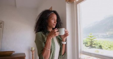 Elinde kahve, pencereden dışarı bakan bir kadın, kendini evinde rahat hisseden. Rahatlama, yaşam tarzı, tefekkür, içecek, rahatlık, sükunet