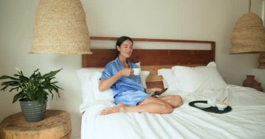 Yatakta dinlenen, mavi pijamalı bir kadın sabah kahvesinin tadını çıkarıyor, evde huzur içinde gülümsüyor. Rahatlama, yaşam tarzı, rahatlık, mutluluk, huzur