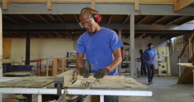 Kulaklık seçiyor, Afro-Amerikalı bir adam tezgah testeresi kullanarak paneli hizalıyor, tam olarak ölçüyorlar. Zanaatkar, atölye, işçilik, kesinlik, güvenlik, ahşap işçiliği, sanayi