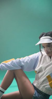 Dikey video: Retro spor giyim giymiş genç bir kadın yeşil arka planda güvenle poz veriyor. Moda, antika, aktivite kıyafeti, tarz, atletik, spor