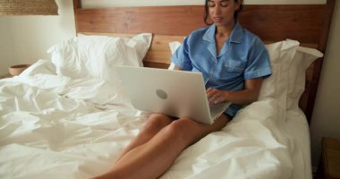 Mavi pijamalı bir kadın yatağında dizüstü bilgisayar kullanıyor, sabah evde dinleniyor. Yaşam tarzı, eğlence, teknoloji, ev ofisi, konfor, sabah rutini