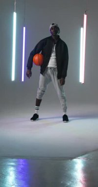Dikey video: Genç adam basketbolu güvenli bir şekilde modern stüdyoda neon ışıklarla tutuyor. Spor, atlet, güven, şehir, moda, dinamik