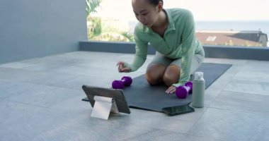 Asyalı kadın çatıdaki egzersiz paspasında diz çökmüş uygulamanın ardından mor dambılları kıvıran tablet tıkırdatıyor. Fitness, wellness, outdoor, lifestyle, modern, health, activity