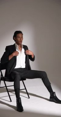 Dikey video: şık takım elbiseli bir adam sandalyede oturuyor, stüdyoda düşünceli görünüyor. Moda, tefekkür, erkek giyim, zarafet, iş, modellik.