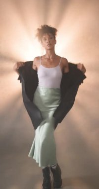 Dikey video: Deri ceketli genç bir kadın yumuşak, dramatik ışıkta rahatça poz veriyor. Moda, portre, tutum, özgüven, heyecan, tarz