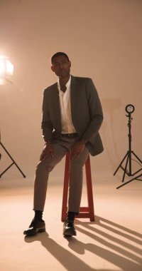 Dikey video: Kendine güvenen şık takım elbiseli adam profesyonel fotoğraf stüdyosunda taburede oturuyor. Moda, güven, erkek giyim, lüks, yakışıklı, yaşam tarzı