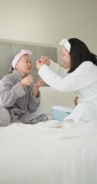 Dikey video: Evde bornozlu iki kadın gülüyor ve yüz maskelerini takıyor. Cilt bakımı, rahatlama, şımartma, bencillik, güzellik, rahatlık.