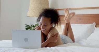 Genç bir kadın laptopunu kullanarak yatakta dinleniyor, evden işine odaklanıyor. Uzaktan çalışma, serbest çalışma, ev ofisi, üretkenlik, teknoloji, konfor