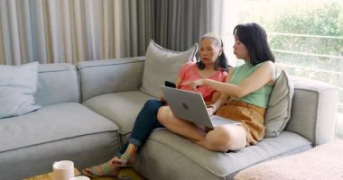 Çinli anne ve kız dizüstü bilgisayar yerleştiriyor, hipodrom kullanıyor, ev kanepesinde ders veriyor. Aile, akıl hocalığı, teknoloji, nesiller arası eğitim, bağlantı, sıcaklık