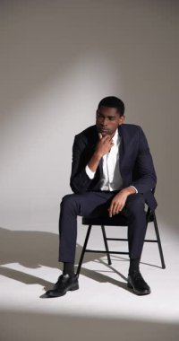 Dikey video: Modern stüdyo ortamında sandalyede oturan şık takım elbiseli kendine güvenen adam. Moda, zarafet, erkek giyim, resmi, iş, profesyonel.