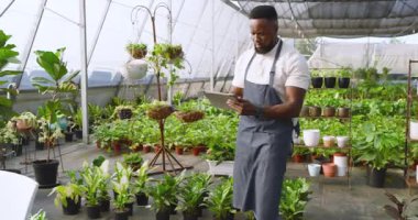 Önlüklü Afro-Amerikan adam serada tablet ve defter kullanıyor. Bahçıvanlık, bahçıvanlık, teknoloji, tarım, ticaret, girişimcilik