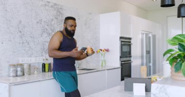Afro-Amerikalı adam şık bir mutfakta duruyor, smoothie içiyor ve akıllı telefondan mesaj atıyor. modern, minimalist, mutfak, akıllı telefon, abur cubur, sağlık, yaşam tarzı