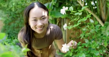 Beyaz çiçekleri fark eden Asyalı kadın bahçedeki çalılıklara yaslanmış yaprakları inceliyor, gülümsüyor. Doğa, dinginlik, kadınlık, açık hava, sükunet, yaşam tarzı, canlı