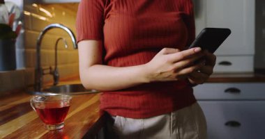 Yetişkin bir Asyalı kadın mutfak tezgahında akıllı telefonları dinliyor, mesaj okuyor ve gülüyor. Gündelik, modern, hayat dolu, yaşam tarzı, refah, evcil, boş zaman