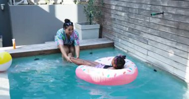 Anne, kızı pembe yüzükle havuz kenarında sallanıyor sonra da kızı yüzdürüyor. Aileler, yaz, rahatlama, boş zaman, açık hava, neşeli, enerjik