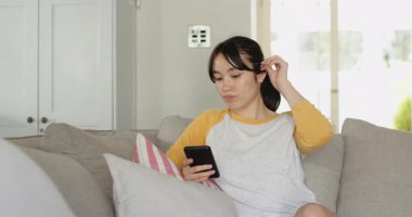 Asian woman scrolling smartphone reacting to content with smile on gray couch in living room. Casual, domestic, contemporary, relaxation, lifestyle, comfort, modern