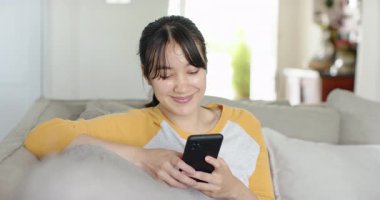 Arkana yaslanıp pencereye bakmadan önce açık gri koltukta akıllı telefonu kaydıran Asyalı kadın. Rahatlık, rahatlık, rahatlık, konfor, yaşam tarzı, iç mekan, modern
